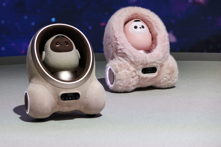 Two small companion robots on a display surface, one seated in a metallic pod and one in a pink furry shell.