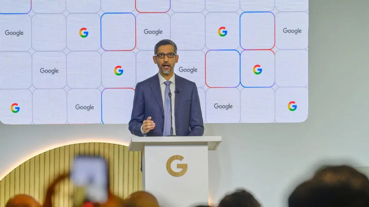 Sundar Pichai speaking at a podium in front of a Google-branded backdrop at a press event.
