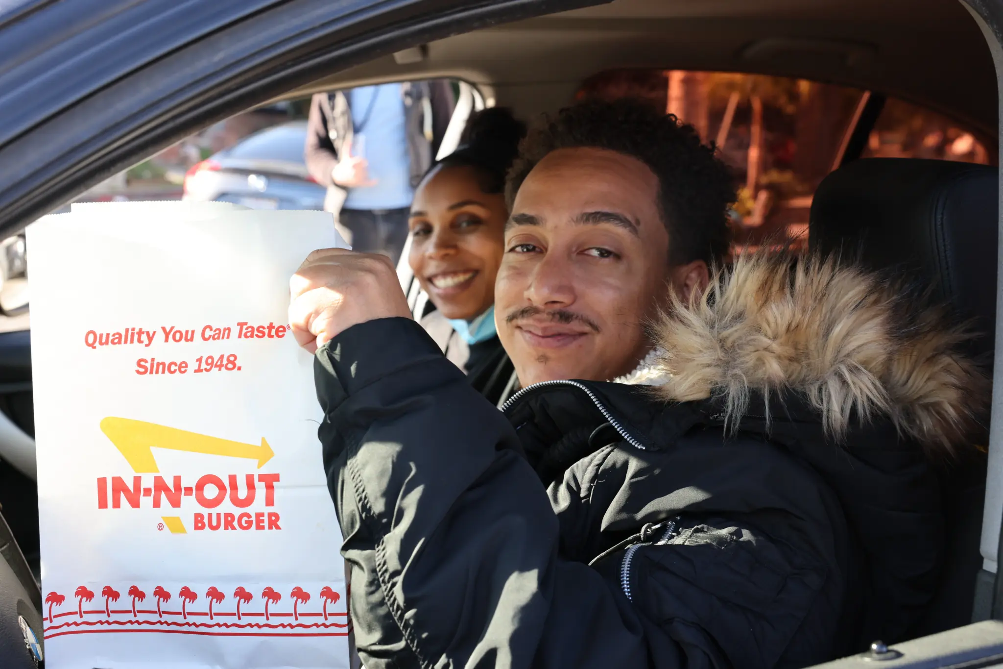 Two people smiling in a car, one holding up a white In-N-Out Burger takeout bag.